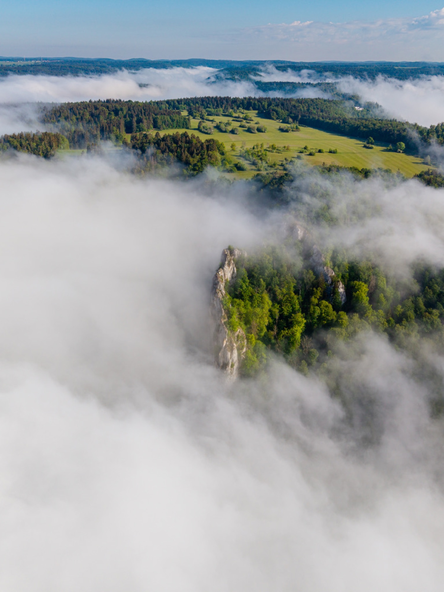 Felsnadel ragt aus dem Nebelmeer, umgeben von grünem Wald