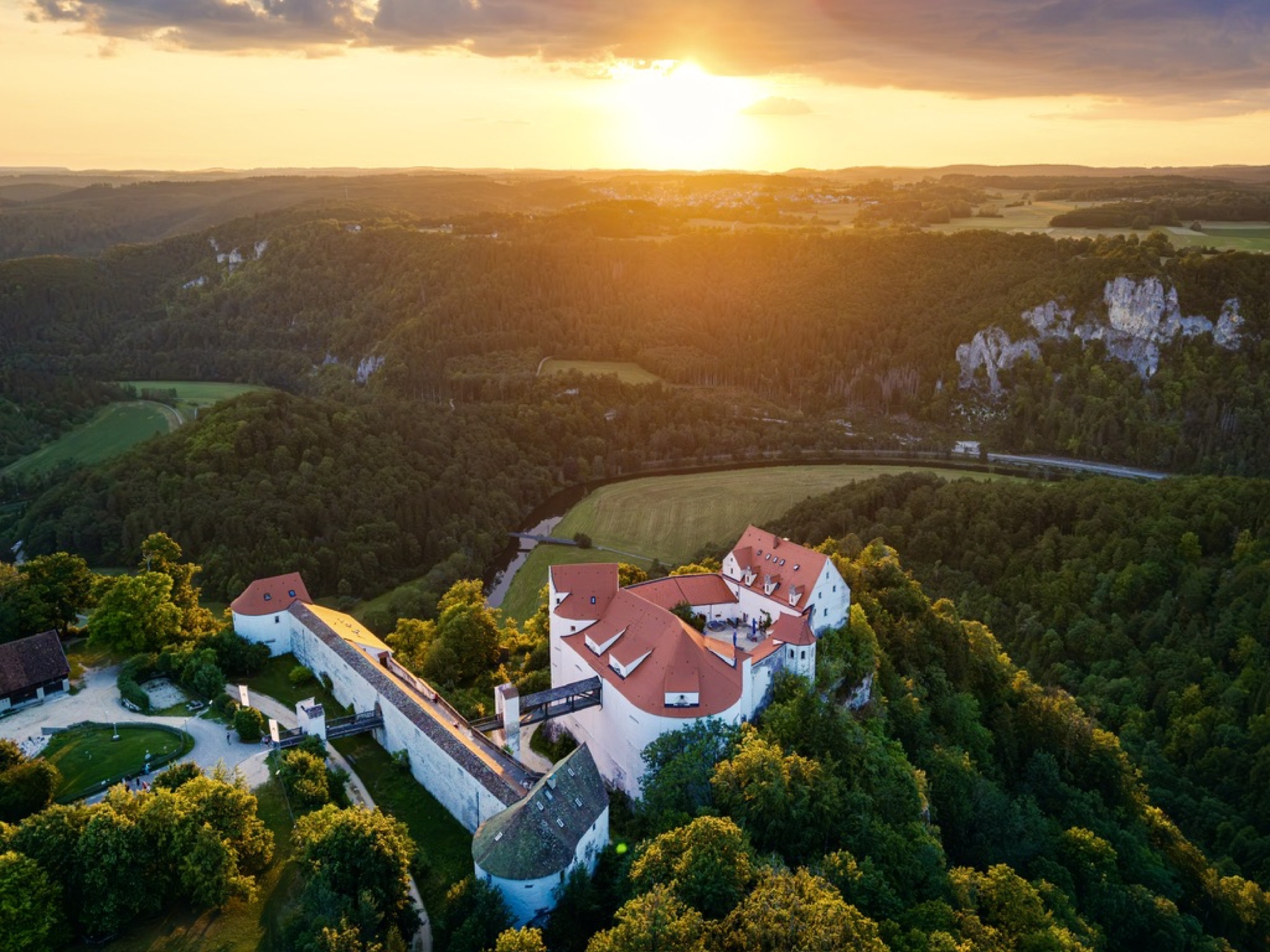Burg auf bewaldetem Hügel im goldenen Abendlicht mit Blick auf einen Flussbogen