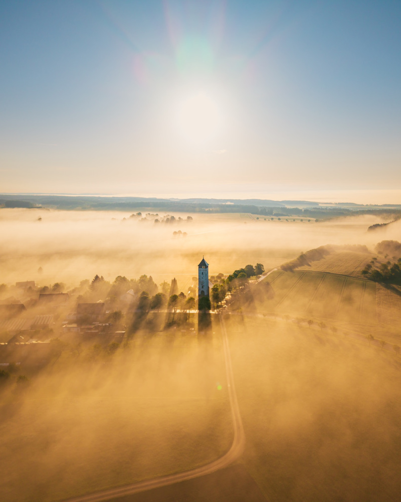 Kirchturm ragt aus goldenem Morgennebel über einem Dorf